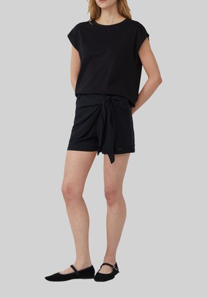 Zwarte korte mouwen top gecombineerd met donkere shorts met een strikdetail aan de voorkant. Model draagt zwarte flats. De textuur lijkt glad en de stof is lichtgewicht.