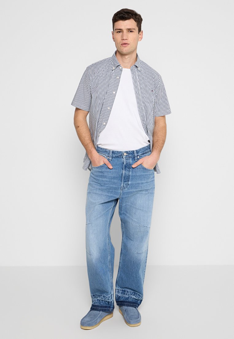 Chemise à manches courtes à rayures noires et blanches sur un T-shirt blanc, associée à un jean en denim baggy bleu clair et des chaussures grises.