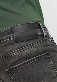 Vaqueros de mezclilla negros con un parche de cuero en la parte trasera que presenta texto en relieve. Incluye dos bolsillos traseros y detalles de costura visibles.