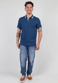 Polo shirt bleu marine avec un col blanc et des accents de manches blancs, associé à un jean bleu clair et des baskets orange. Design simple et décontracté.