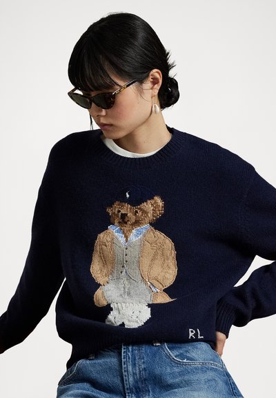Navy-blauw gebreid sweater met een geborduurd beer in een blazer, met een witte shirt en blauwe stropdas. Inclusief logo "RL" onderaan.