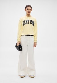 Sárga pulóver "GANT 1949" nyomattal, fehér bőszárú nadrág, fekete kézitáska és ezüst platform cipők minimalista környezetben.