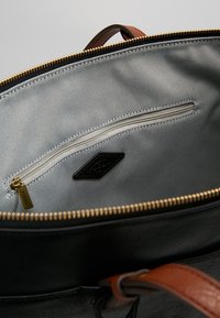 Zwarte leren tas interieur met een gladde grijze stofvoering, voorzien van een ritszak en een bruine leren handgreep. Gouden rits zichtbaar.