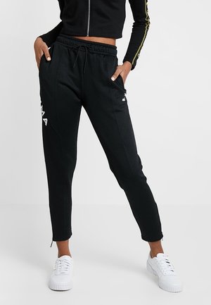 Personne portant un pantalon de jogging noir avec taille à cordon et des baskets blanches, debout les mains dans les poches devant un fond uni.