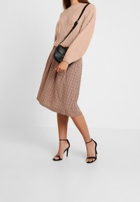 Pull beige en maille à manches longues, associé à une jupe midi à carreaux dans des tons de marron et rouge, et des sandales à talons noires avec des lanières.