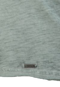 T-shirt di cotone verde chiaro con una texture mélange e un piccolo tag metallico che dice "Key Largo" nell'angolo in basso a sinistra.