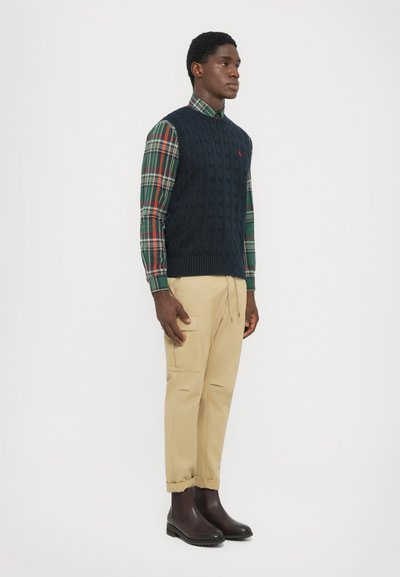Hombre vistiendo un chaleco de punto azul marino sobre una camisa a cuadros verde y roja, pantalones cargo beige y botas de cuero marrón oscuro, posando contra un fondo blanco.
