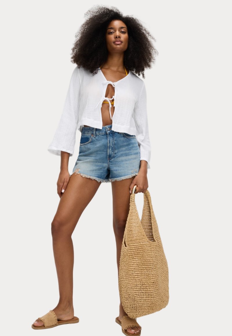 Blusa bianca corta con nodo frontale, pantaloni corti di denim e borsa a tracolla intrecciata marrone. Il modello indossa sandali scivoli.