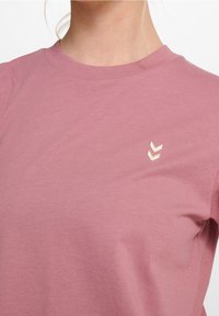 Kurzärmliges T-Shirt aus softem rosa Baumwollstoff, mit Rundhalsausschnitt und einem kleinen cremefarbenen Chevron-Logo im linken Brustbereich.