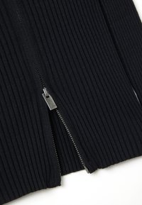 Tejido negro con textura acanalada, que presenta un cierre central con herrajes plateados y muestra rayas verticales texturizadas. Los acabados de los bordes son suaves.