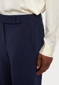 Marineblauwe broek van structuurstof met een vlakke voorkant, riemlussen en zijzakken, gecombineerd met een crèmekleurig shirt met lange mouwen.