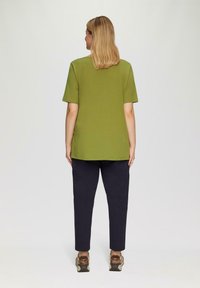 Vrouw met schouderlang blond haar, gekleed in een groene kortemouwenshirt, zwarte broek en bruine sneakers, staat met haar rug naar de camera gekeerd.