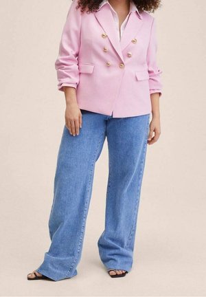 Persoon draagt een roze double-breasted blazer met gouden knopen, lichtblauwe wijd uitlopende jeans en zwarte open sandalen.