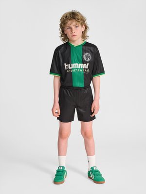 Boy with curly hair stands wearing a black and green Hummel sportswear jersey, black shorts, white socks, and green sneakers on gray background.