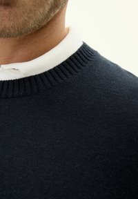 Mörkblå stickad tröja med en ribbad rundhalsad krage och en vit innerkrage, som visar upp en mjuk textur och en klassisk design.