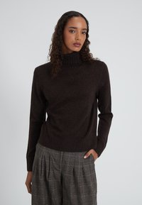 Donna con capelli ricci che indossa un pullover a collo alto marrone scuro e pantaloni a pieghe grigi a quadri, in piedi con una mano in tasca.