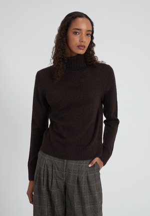 Frau mit lockigem Haar, die einen dunkelbraunen Rollkragenpullover und graue kariertes, gefältete Hose trägt, steht mit einer Hand in der Tasche.