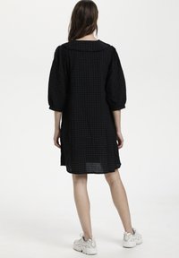 Vestido de cuadros negros con cuello redondo, mangas abullonadas tres cuartos y un corte holgado, combinado con zapatillas de color claro.
