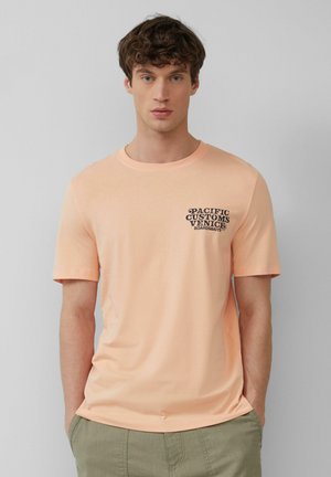 Perzikkleurig katoenen T-shirt met korte mouwen, ronde halslijn en zwarte bedrukt tekst op de linkerborst met de tekst "PACIFIC CUSTOMS VENICE BOARDS&SK8S."