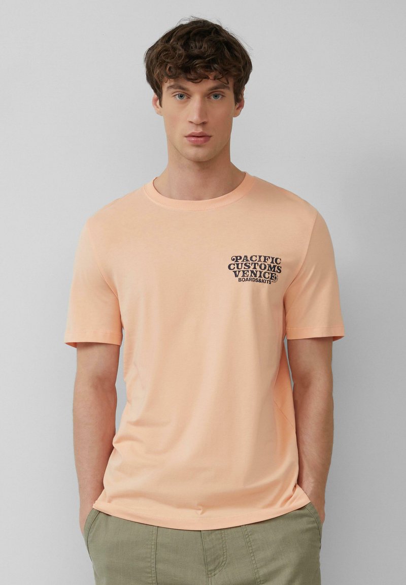 Perzikkleurig katoenen T-shirt met korte mouwen, ronde halslijn en zwarte bedrukt tekst op de linkerborst met de tekst "PACIFIC CUSTOMS VENICE BOARDS&SK8S."