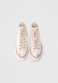 Λευκά ψηλά αθλητικ ά παπούτσια από καμβά με κορδόνια διακοσμημένα με κόκκινη καρδιά, κόκκινη λαστιχένια σόλα και λεπτομέρεια καρδιάς στο δάχτυλο.