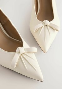Bailarinas blancas de punta fina, hechas de cuero suave, con un lazo decorativo y pliegues en la punta, un interior sencillo y mínimos detalles.