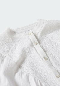 Weiße bestickte Bluse mit Rundhalsausschnitt, Knopfverschluss, strukturiertem Stoff und komplexen Mustern an der Vorderseite und den Ärmeln.