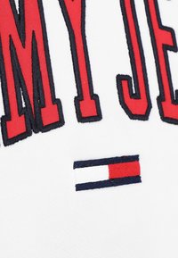 Weiße Stoff mit großen roten gestickten Buchstaben "MY JE", umrandet in Marineblau. Verfügt über ein kleines rechteckiges Logo-Patch in Marineblau, Weiß und Rot.