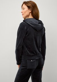 Survêtement en velours gris foncé comprenant une veste à capuche au toucher lisse et un pantalon assorti avec une poche arrière et un détail à pression.