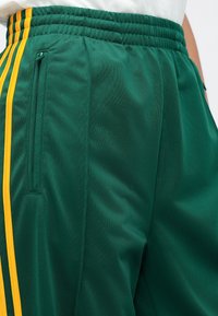 Zielone sportowe szorty o teksturowanej powierzchni, z żółtymi paskami po bokach i kieszenią na zamek na prawym biodrze. Elastyczny pas.