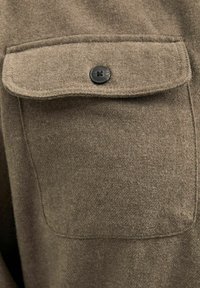 Camisa de tela texturizada marrón con un bolsillo en el pecho que tiene un botón negro y detalles de costura visibles a lo largo de las costuras.