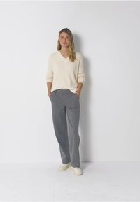 Krémový svetr s výstřihem do V a dlouhými rukávy, v kombinaci se šedými kalhotami s širokými nohavicemi a pruhy. Model má na sobě bílé tenisky a ruce má v kapsách.