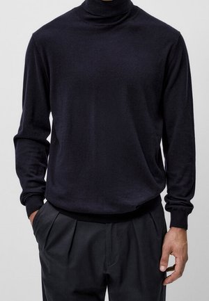 Homme portant un pull noir ajusté à col roulé et un pantalon noir plissé, une main dans la poche et l'autre détendue.