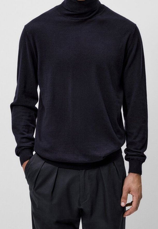 Homme portant un pull noir ajusté à col roulé et un pantalon noir plissé, une main dans la poche et l'autre détendue.