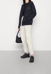 Sweat à capuche marine avec logo, haut noir, joggeurs crème et baskets noires. Petit sac noir, design décontracté, coupe ample et poignets côtelés.
