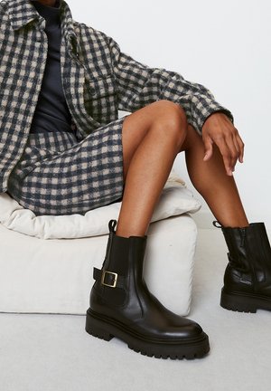 Person sitting with crossed legs wearing checked jacket and skirt, black ankle boots with buckle, on a light-colored cushion.