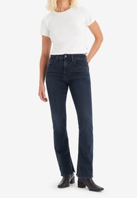 Jeans oscuros de tiro alto con corte recto y aberturas laterales, combinados con una camiseta blanca ajustada y botines negros.