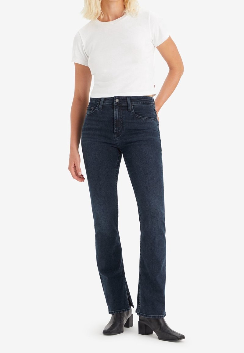 Jeans oscuros de tiro alto con corte recto y aberturas laterales, combinados con una camiseta blanca ajustada y botines negros.