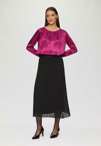 Magenta satynowa bluzka z długimi rękawami i plisami, w połączeniu z czarną plisowaną spódnicą. Modelka ma na sobie czarne szpilki. Neutralne tło.