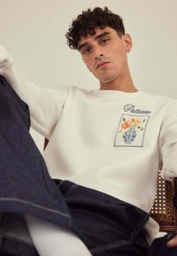Sweatshirt blanc avec un patch à motif floral sur la poitrine ; associé à un pantalon en denim bleu foncé. Tissu texturé, coupe décontractée, détails minimalistes.