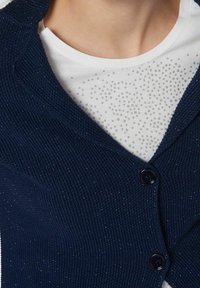 Cardigan lavorato a maglia blu navy con un motivo testurizzato, caratterizzato da un collo a V, due bottoni e una camicia bianca sotto con dettagli argentati a pois.
