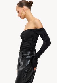 Vrouw met haar in een knot draagt grote gouden oorbellen, een ruche off-shoulder langemouwen zwart top en een hoog gewaardeerde zwarte leren rok.