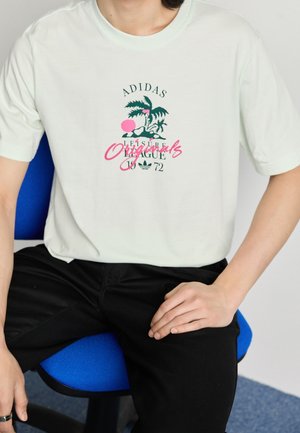 Person sitzt auf einem blauen Stuhl und trägt ein weißes Adidas Originals T-Shirt mit einem Palmbaum-Graphic und schwarze Hose.