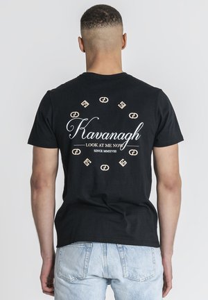 Mężczyzna zwrócony plecami, ubrany w czarną koszulkę z tekstem i symbolami "Kavanagh Look At Me Now Since MMXVIII" w okręgu na plecach.