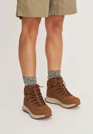 Benedenbenen met beige korte broek, grijze gebreide sokken en bruine veterschoenen voor wandelen met lichtgekleurde zolen op een effen achtergrond.