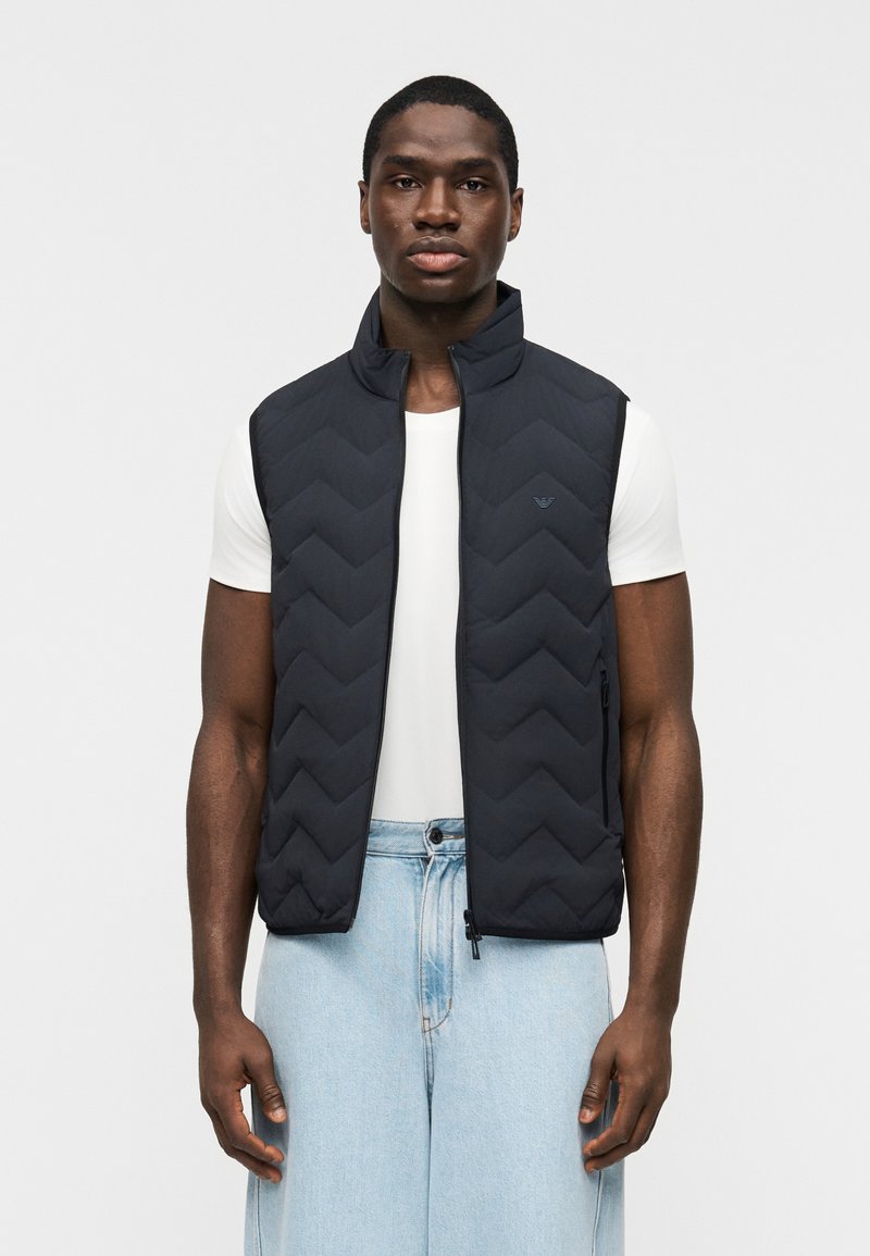 Άνδρας που φοράει μαύρο γιλέκο με φερμουάρ και επένδυση πάνω από λευκό πουκάμισο με κοντά μανίκια και ανοιχτό μπλε τζιν, στέκεται μπροστά σε λευκό απλό φόντο.