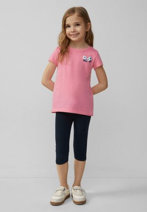 Roze katoenen T-shirt met korte mouwen, versierd met geborduurde bloemen, gecombineerd met donkergroene leggings en witte sneakers met bruine accenten.