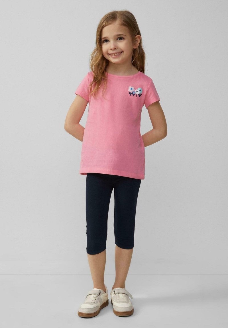 T-shirt en coton rose à manches courtes, avec des fleurs brodées, associé à des leggings bleu foncé et des baskets blanches avec des accents marron.