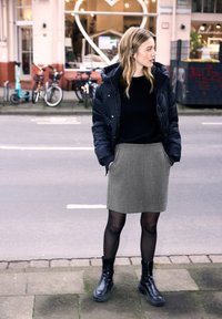 Giacca a vento nera con cappuccio, maglione nero, gonna grigia con tasche, collant neri e stivali neri chunky; in piedi su un marciapiede.