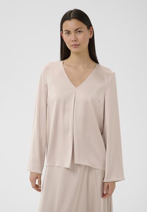 Egyenes sötét hajú nő világos bézs színű V-nyakú, hosszú ujjú blúzt és hozzáillő szoknyát visel egyszerű háttér előtt.